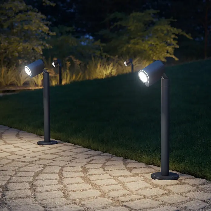 Steinel Lámpara de pie para exterior Spot Way - Color del marco: Antracita, Amarillo, 22,6 x 14 x 57,2 cm, Con bombillas - Escena de jardín con dos luces de bolardo que iluminan el camino, con luces adicionales al fondo que crean un ambiente atmosférico.