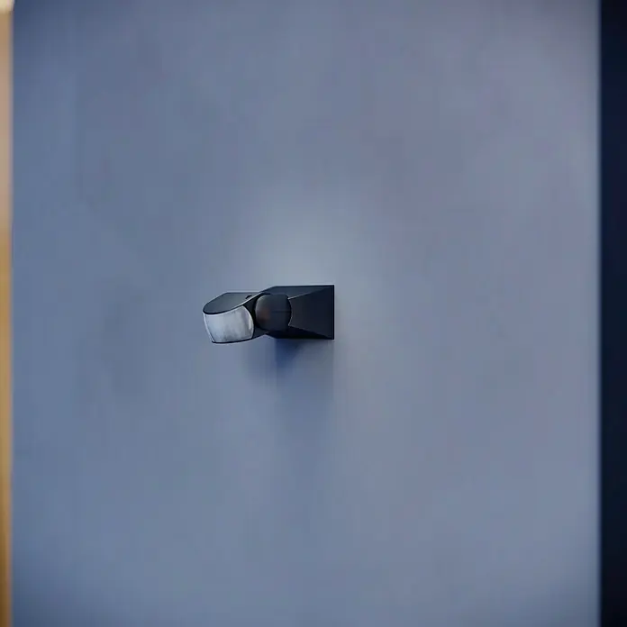 Steinel Sensor de movimiento IS 1 - Alcance sensor: 10 m, Negro, Ángulo de detección sensor: 120 °, L x An x Al: 120 x 80 x 50 mm - La imagen muestra un detector de movimiento negro moderno montado en una pared azul, diseñado para activar la iluminación automáticamente.
