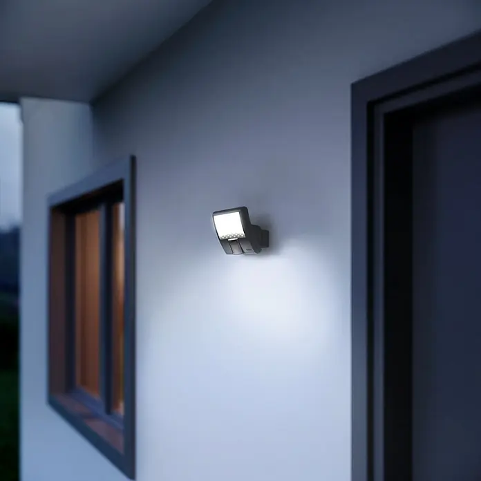 Steinel LED-Sensor-Strahler XLED home Curved S anthrazit - 9,3 W, L x B x H: 16 x 10,7 x 12 cm, Anthrazit, IP44 - Moderne Außenleuchte an einer weißen Wand. Die Lampe strahlt helles, weißes Licht aus und sorgt für Sicherheit und Ambiente im Außenbereich.
