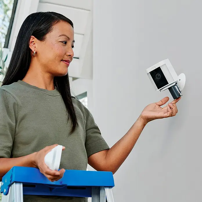 Ring Überwachungskamera Spotlight Cam Plus Battery - Batteriebetrieben, 1.080 Pixel (Full HD), Weiß, B x H: 7,6 x 12,6 cm, Akku - Eine Frau montiert eine Ring-Überwachungskamera an einer Wand, wobei die Kamera und ihre Installationsbasis gut sichtbar sind.