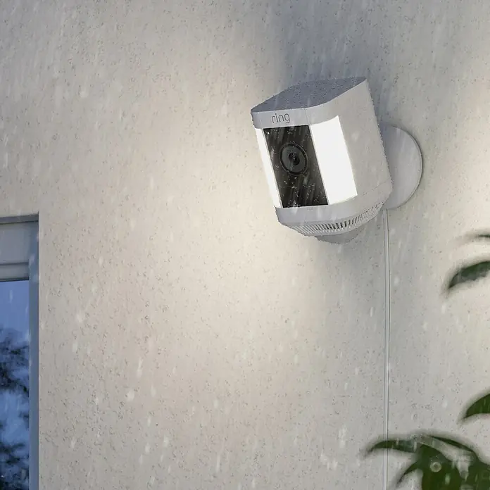 Ring Überwachungskamera Spotlight Cam Plus Plugin - Netzbetrieben, 1.080 Pixel (Full HD), Weiß, B x H: 7,6 x 12,6 cm, Netzanschluss - Eine Ring Spotlight Cam ist an einer weißen Wand montiert und wird im Freien bei Regenwetter gezeigt.