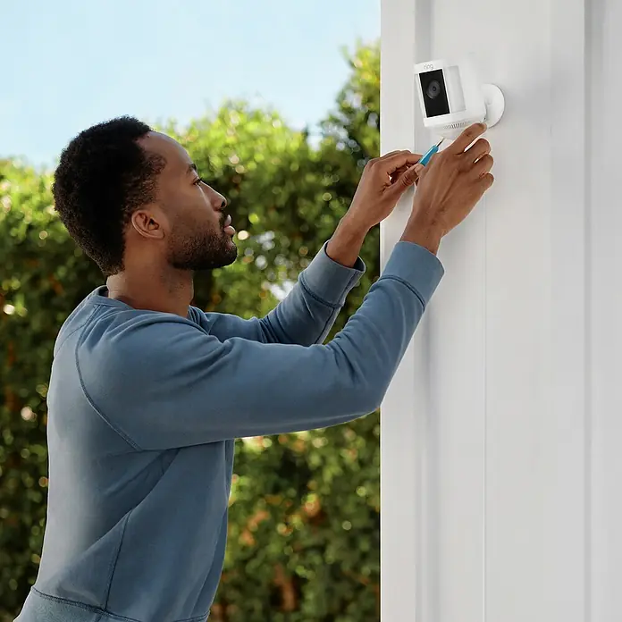Ring Überwachungskamera Spotlight Cam Plus Plugin - Netzbetrieben, 1.080 Pixel (Full HD), Weiß, B x H: 7,6 x 12,6 cm, Netzanschluss - Ein Mann befestigt eine Ring-Überwachungskamera an einer weißen Wand im Freien mit einem Schraubenzieher.