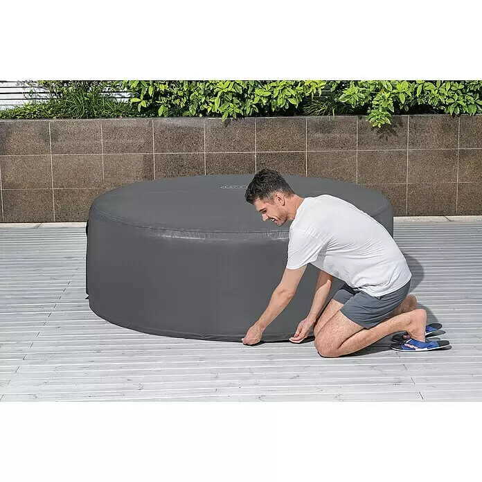 Bestway Lay-Z-Spa Pool-Abdeckplane - Passend für: Runde Whirlpools bis Ø x H: 180 x 66 cm, Ø x H: 180 x 66 cm - Ein Mann kniet auf einer Terrasse und passt die graue Abdeckung eines runden aufblasbaren Whirlpools an.