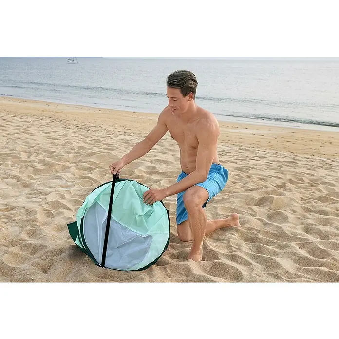 Bestway Pavillo Pop-Up StrandmuschelL x B x H: 200 x 120 x 90 cm, Türkis - Ein Mann kniet am Strand und öffnet einen grünen und weißen, runden Strand-Pop-Up-Unterstand, um sich vor Sonne und Wind zu schützen.