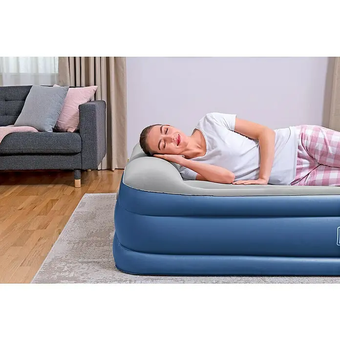 Bestway Luftbett Vento - L x B x H: 203 x 152 x 46 cm, Integrierte Luftpumpe - Eine Frau schläft entspannt auf einer blauen Luftmatratze mit integriertem Kopfkissen in einem gemütlichen Raum mit Sofa und Vorhängen.