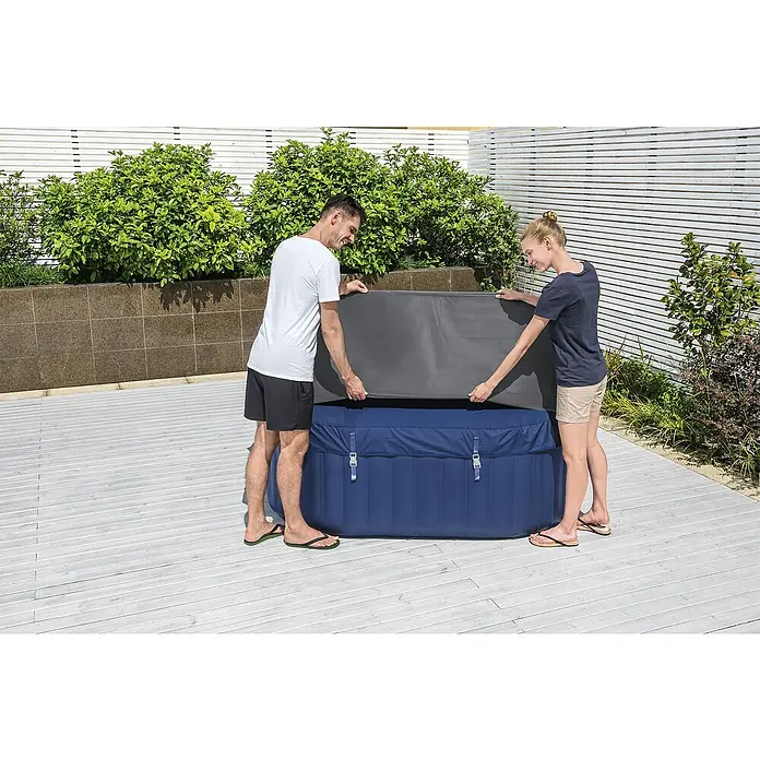 Bestway Lay-Z-Spa Pool-AbdeckplanePassend für: Quadratische Whirlpools bis L x B x H: 180 x 180 x 71 cm, L x B x H: 180 x 180 x 71 cm - Zwei Personen decken einen blauen, aufblasbaren Whirlpool im Freien mit einer grauen Abdeckung ab.
