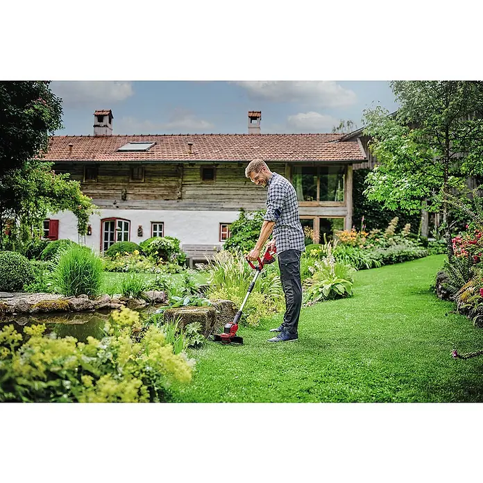 Einhell PXC 18V Akku-Rasentrimmer GE-CT 18/28 Li-Solo - 18 V, 5.200 mAh, Ohne Akku, Schnittbreite Messer: 28 cm, Schnittbreite Faden: 28 cm - Ein Mann benutzt einen Einhell Akku-Rasentrimmer im Garten, umgeben von üppigem Grün und einem charmanten Haus im Hintergrund.
