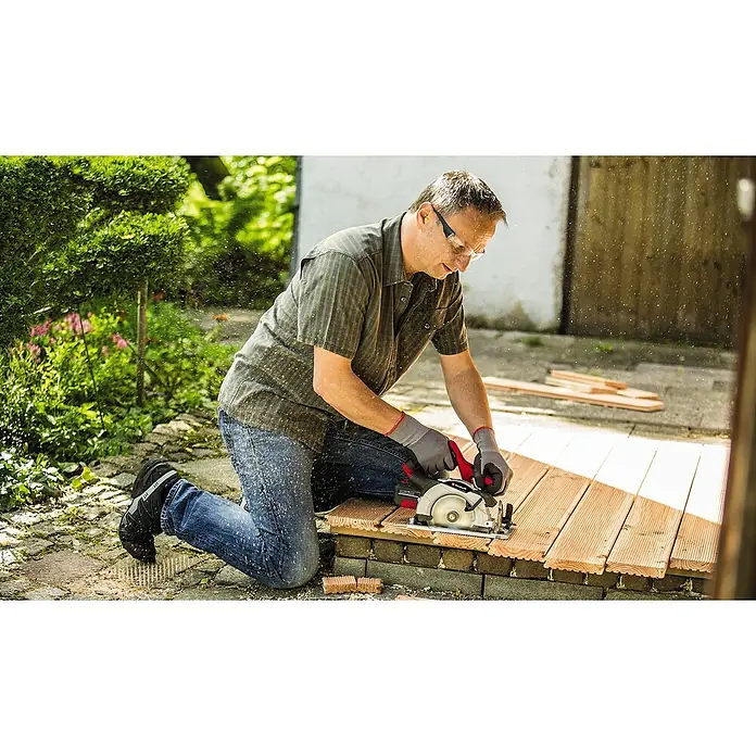 Einhell Power X-Change 18V Akku-Handkreissäge TE-CS 18/165-1 Li - Solo - 18 V, Ohne Akku, Leerlaufdrehzahl: 4.200 U/min - Ein Mann kniet und schneidet mit einer Einhell Kreissäge ein Holzbrett im Freien, umgeben von Werkzeugen und Holzschnitzeln.