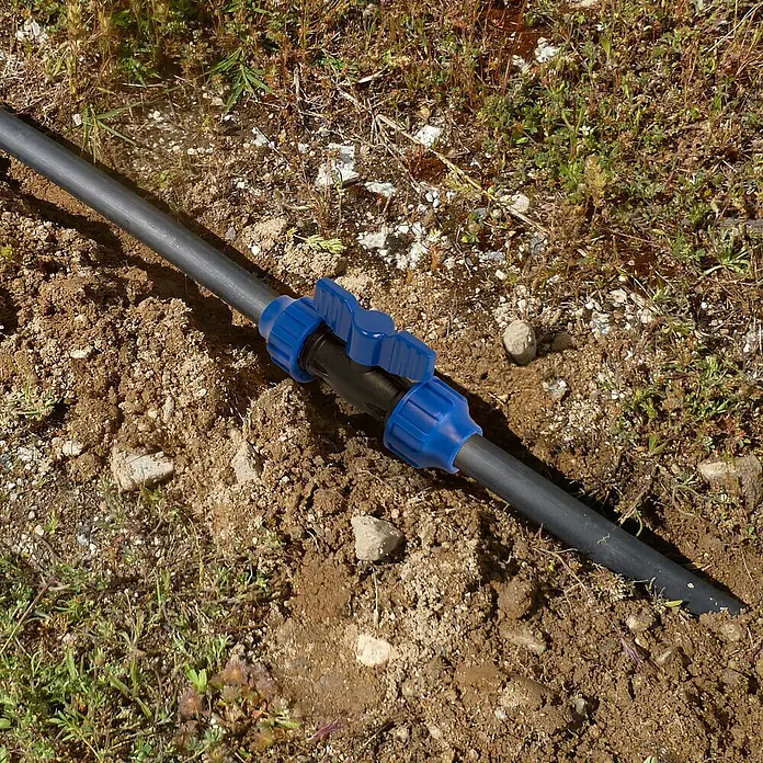 Válvula esférica PE para riego - Diámetro: 20 mm, Polietileno - Primer plano de un tubo de riego negro con válvula de cierre azul, enclavado en la tierra y rodeado de escasa vegetación.