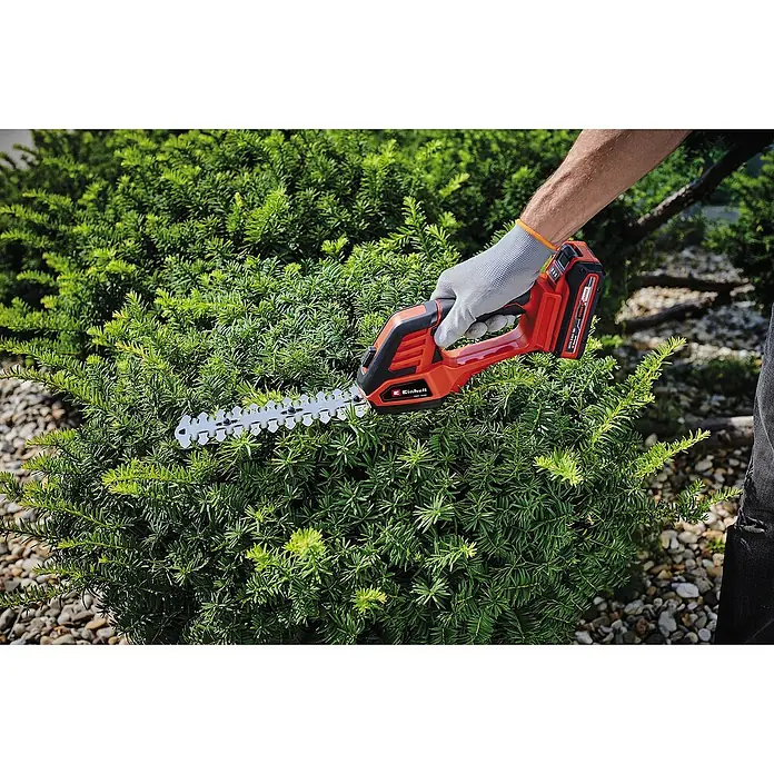 Einhell Power X-Change 18V Akku-Grasschere CE-CG18/100 LI T-SOLO - 18 V, Ohne Akku, Schnittbreite: 100 mm - Eine rote Einhell Akku-Heckenschere beim Trimmen einer grünen Hecke, in der Hand eines Gärtners mit grauem Handschuh.