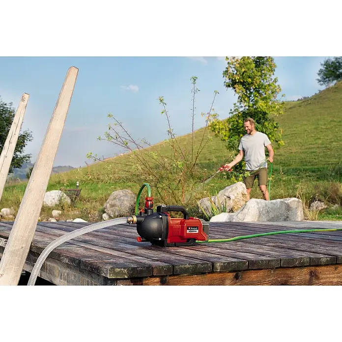 Einhell Power X-Change 18V Akku-Gartenpumpe AQUINNA 36/30 - Ohne Akku, Max. Fördermenge: 3.000 l/h - Eine rote Einhell Aquinna Gartenpumpe mit schwarzem Griff und Schläuchen liegt auf einer hölzernen Plattform. Im Hintergrund gießt ein Mann.