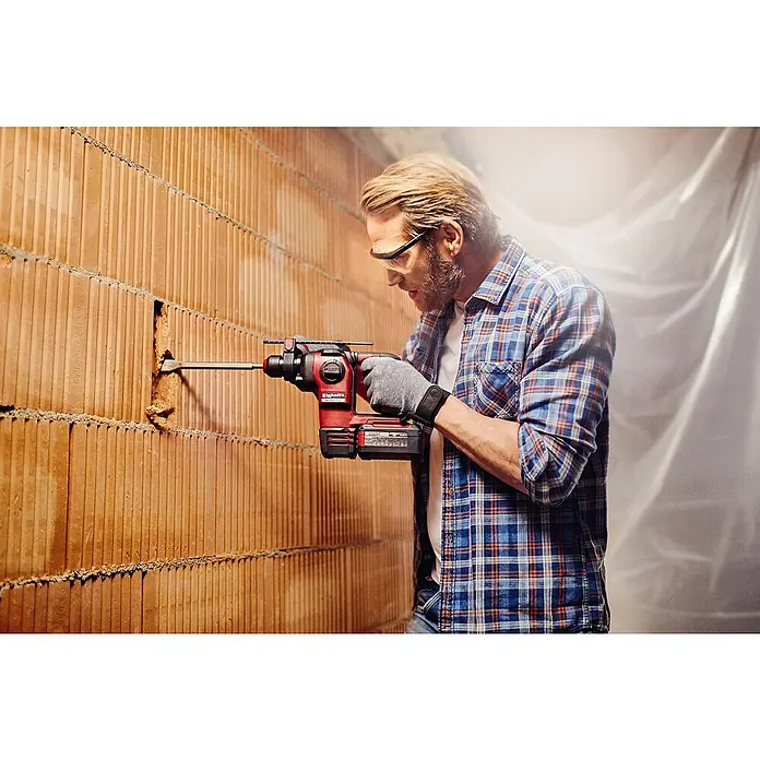 Einhell Professional PXC 18V Martillo combinado de batería Herocco - 18 V, Sin batería, 0 Ah, 2,2 J - Un hombre perfora una pared de ladrillos con un martillo rotatorio Einhell, incluidas gafas de seguridad y guantes.
