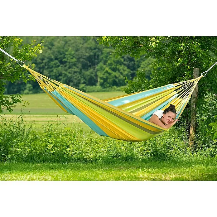 Amazonas Doppelhängematte Colombiana - L x B: 350 x 160 cm, Limona - Eine Frau entspannt in einer bunten Hängematte im Garten, die zwischen zwei Bäumen aufgehängt ist und Ruhe ausstrahlt.