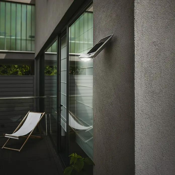 Lutec Aplique solar para exterior con sensor Drop - 2,3 W, L x An x Al: 19 x 21 x 22 cm, Plateado, IP44 - Aplique de pared metálico moderno con panel solar, que emite una luz brillante sobre un patio. El diseño es minimalista y funcional.