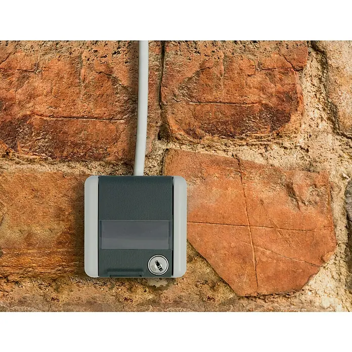 Jung WG 800 Feuchtraum-Steckdose 820NAWSL - Anthrazit/Grau, 1-fach, Aufputzmontage, IP44 - Nahaufnahme einer grauen Schlüsselbox mit Kabel an einer strukturierten Mauer. Ideal zur sicheren Schlüsselaufbewahrung im Außenbereich.