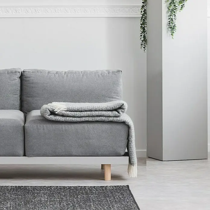 Rei Pata para muebles - L x An x Al: 5,8 x 5,8 x 15 cm, Capacidad de carga: 20 kg, Madera - Primer plano de un sofá gris con una manta doblada y una pequeña mesa de madera, sobre una pared blanca lisa.