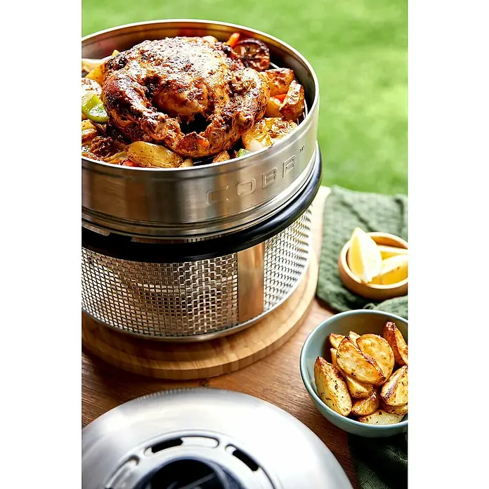 Cobb Hähnchen-Bräter mit Deckelerhöhung - Edelstahl, Passend für: Cobb Grillmodelle / Cobb Gasgrill Deluxe / Cobb Holzkohlegrill Air Deluxe - Ein Cobb Grill mit einem gebratenen Hähnchen und Kartoffeln, serviert auf einem Holztisch mit einem grünen Tuch.