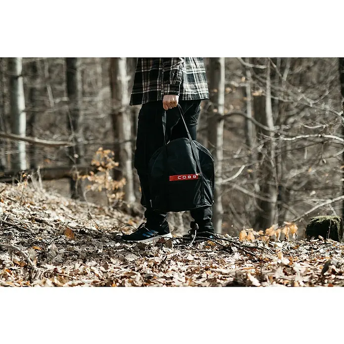 Cobb Transporttasche - Textil, Passend für: Cobb Holzkohlegrill Air Deluxe - Eine Person im Wald hält eine schwarze Cobb-Tasche mit rotem Logo. Ideal für Outdoor-Abenteuer, robust und stilvoll.