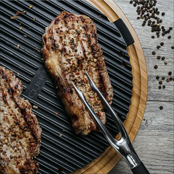 Cobb Grillplatte Griddle+Aluminium, Durchmesser: 27 cm, Passend für: Cobb Grillmodelle / Cobb Gasgrill Deluxe / Cobb Holzkohlegrill Air Deluxe - Gegrilltes Steak mit Gewürzen auf einer Grillplatte, gehalten mit einer Grillzange.