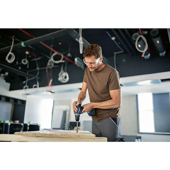 Bosch Professional AMPShare 18V Akku-Bohrschrauber GSR 18V-55 - 18 V, 2 Akkus, 4 Ah - Ein Handwerker bohrt mit einer Bosch Bohrmaschine ein Loch in ein Holzbrett, um Bauarbeiten durchzuführen. Schutzbrille für Sicherheit.
