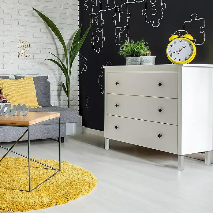 Rei Möbelfuß - L x B x H: 4 x 8 x 10 cm, Kunststoff, Aluminium, 1 Stk. - Helle Kommode mit drei Schubladen, ideal für modernes Interieur, ergänzt durch eine gelbe Uhr und grüne Pflanzen auf der Oberfläche.