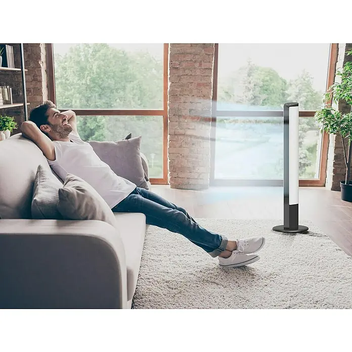 Purline Venty Ventilador de torre TO 55 - Blanco y Negro, 113 cm - Un hombre se relaja en el sofá junto a un purificador de aire elegante y moderno con un diseño minimalista en un salón luminoso.