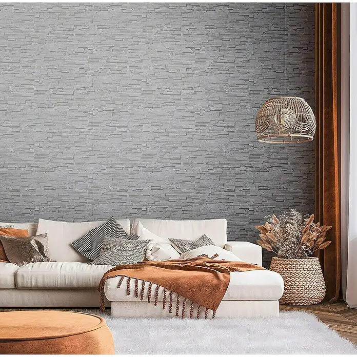 A.S. Création Metropolitan Stories Travel Styles Vliestapete - Anthrazit, Steinoptik, 10,05 x 0,53 m - Wohnzimmer mit weißem Sofa, Zierkissen und dekorativer Lampe vor einer Wand mit Steinmuster, die eine warme und einladende Atmosphäre schafft.