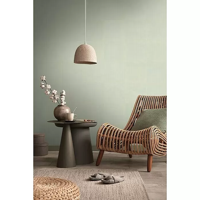 A.S. Création Metropolitan Stories Travel Styles Vliestapete - Sandgrün, Uni, 10,05 x 0,53 m - Ein gemütlicher Wohnbereich mit einem geflochtenen Rattansessel, einem runden Jute-Pouf und einer Seil-Pendelleuchte, die eine natürliche und entspannte Atmosphäre schaffen.