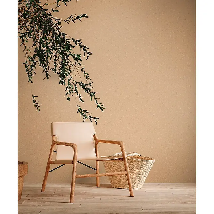 A.S. Création Metropolitan Stories Travel Styles VliestapeteMelange-Orange, Uni, 10,05 x 0,53 m - Ein beiger Ledersessel aus Holz steht neben einem Strohkorb vor einer schlichten Wand mit Blattwerk.