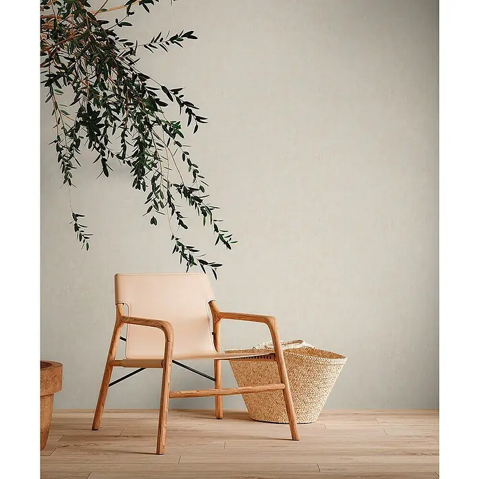 A.S. Création Metropolitan Stories Travel Styles VliestapeteHellbeige, Uni, 10,05 x 0,53 m, Spachtelstruktur - Elegante Lederstuhl mit Holzrahmen und Strohkorb, vor einer sanften Wand mit grünen Zweigen, für eine stilvolle Inneneinrichtung.