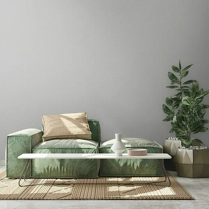 A.S. Création Metropolitan Stories Travel Styles Vliestapete - Melange-Grau, Uni, 10,05 x 0,53 m - Gemütliches grünes Sofa mit Kissen und Tisch, neben einer Topfpflanze auf einem geflochtenen Teppich.