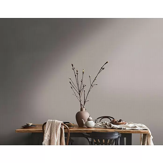 A.S. Création Metropolitan Stories Travel Styles Vliestapete - Taupe, Uni, 10,05 x 0,53 m - Texturierte Wand mit vertikalem Farbverlauf von dunklem Graubeige zu hellem Beige, als Kulisse für einen Holzesstisch.
