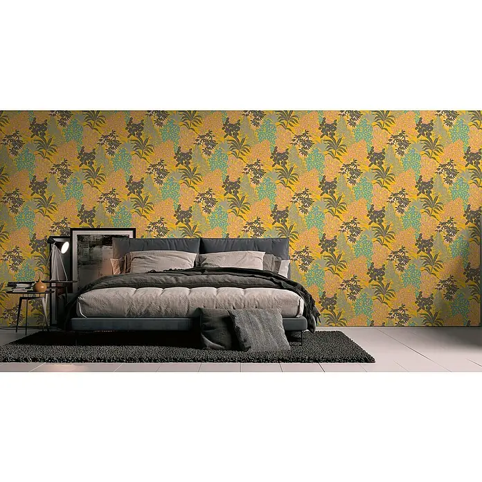 A.S. Création Metropolitan Stories Travel Styles Vliestapete Blumen I - Gelb, Floral, 10,05 x 0,53 m - Ein Schlafzimmer mit einer floralen Tapete in Gelb-, Grau-, Rosa- und Blautönen, einem grauen Bett und einem dunklen Teppich.