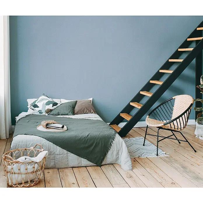 A.S. Création Metropolitan Stories Travel Styles VliestapeteMelange-Blau, Uni, 10,05 x 0,53 m - Ein gemütliches Schlafzimmer mit einem Bett mit grüner Decke, einer Leiter und einem Korbsessel, das eine entspannende Atmosphäre schafft.