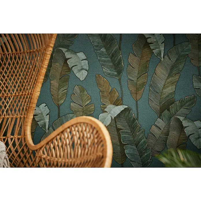 A.S. Création Metropolitan Stories Travel Styles Vliestapete - Blau/Gold, Floral, 10,05 x 0,53 m - Nahaufnahme eines Rattan-Stuhls vor einer Wand mit tropischem Blättermuster, das eine exotische und entspannende Atmosphäre schafft.