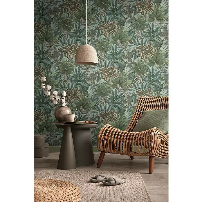 A.S. Création Metropolitan Stories Travel Styles Vliestapete - Grün/Beige, Floral, 10,05 x 0,53 m - Wohnzimmer-Szene mit einem Rattan-Sessel, einem Beistelltisch und einer Hängelampe vor einer grünen Blättertapete.