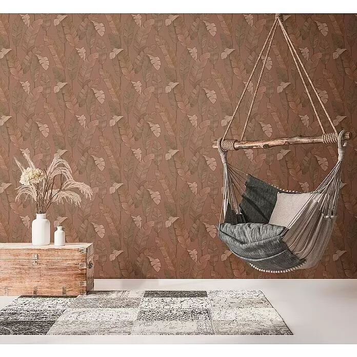 A.S. Création Metropolitan Stories Travel Styles Vliestapete - Rost, Floral, 10,05 x 0,53 m - Terrakotta-Tapete mit tropischem Blättermuster, die eine gemütliche Ecke mit Hängesessel und rustikaler Holztruhe schmückt.