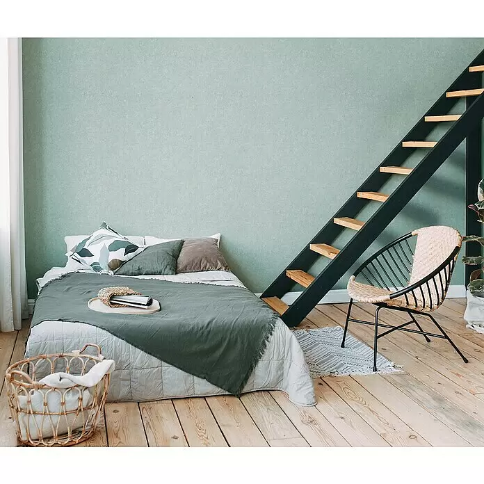 A.S. Création Metropolitan Stories Travel Styles Vliestapete - Hellgrün, Uni, 10,05 x 0,53 m, Feine Struktur - Ein stilvolles Schlafzimmer mit einem Bett in Grüntönen, einem Stuhl aus Naturgeflecht und schwarzem Metall sowie einer schwarzen Holztreppe zum Loft.
