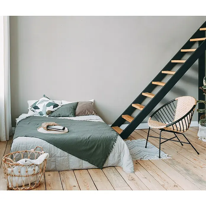 A.S. Création Metropolitan Stories Travel Styles VliestapeteWeiß, Uni, 10,05 x 0,53 m - Schlafzimmer mit einem Doppelbett, einer Leiter an der Wand und einem Korbstuhl auf einem hellen Holzboden. Die Bettwäsche ist in Grüntönen gehalten.
