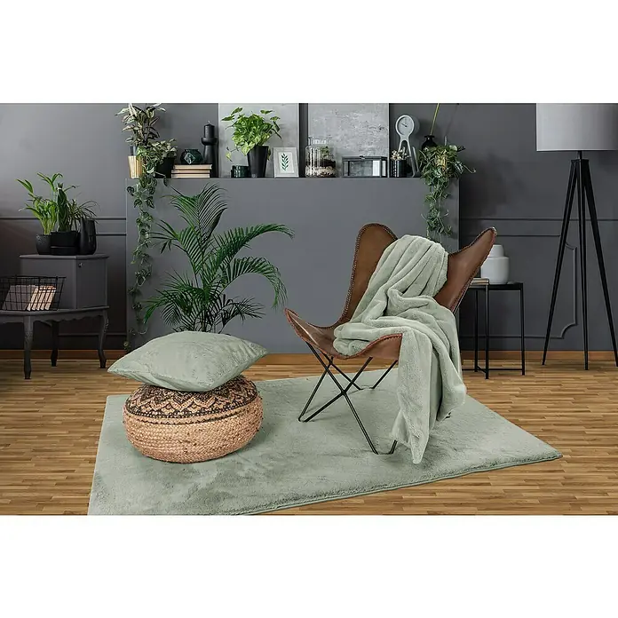Decke Happy - Jade, 200 x 150 cm, 100 % Polyester - Ein gemütliches Wohnzimmer mit einem braunen Sessel, einer weichen grünen Decke, einem Kissen und einem Pouf auf einem flauschigen grünen Teppich.