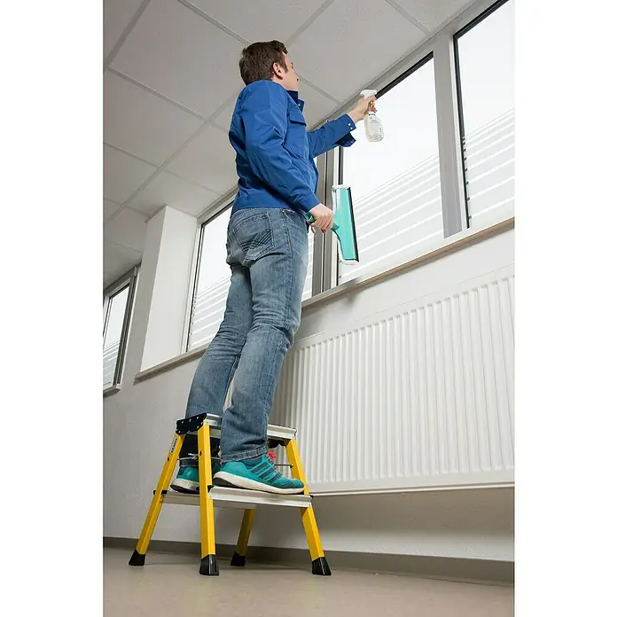 Krause Monto Klapptritt Rolly - Anzahl Tritte: 4 Stk., Arbeitshöhe: 2,45 m, Belastbarkeit: 150 kg, Aluminium, Gelb - Mann auf einer Krause-Leiter beim Fensterputzen mit Sprühflasche und Abzieher in einem Büro.