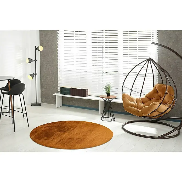 Fellteppich Happy - Amber, Durchmesser: 160 cm, 100 % Polyester (Flor) - Ein runder, brauner Teppich liegt in einem modern eingerichteten Raum mit einem Hängesessel und einer Stehlampe. Der Teppich sorgt für Wärme und Komfort.