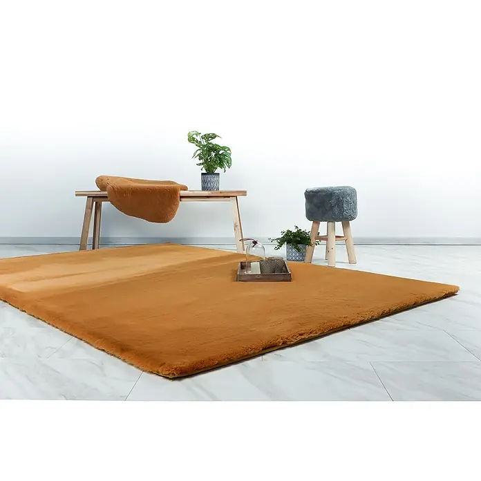 Fellteppich Happy - Amber, 230 x 160 cm, 100 % Polyester (Flor) - Flauschiger, senffarbener Teppich auf einem Marmorboden, mit Beistelltisch, Hocker und Pflanzen.