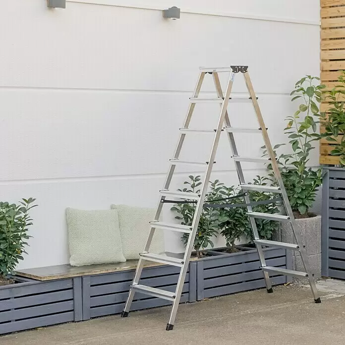 Krause Monto Doppel-Stehleiter Dopplo - Arbeitshöhe: 3,3 m, Anzahl Tritte: 16 Stk., Belastbarkeit: 150 kg, Aluminium - Silberne Aluminium-Klappleiter steht draußen neben einer Wand und Pflanzen.