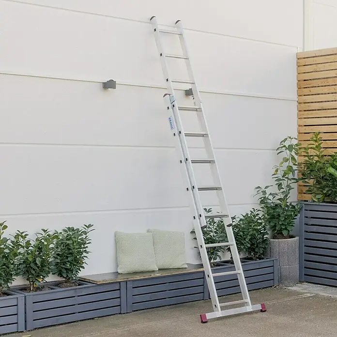 Krause Corda Schiebeleiter - Anzahl Tritte: 16 Stk., Arbeitshöhe: 4,95 m, Belastbarkeit: 150 kg, Aluminium - Eine silberne Mehrzweckleiter von Krause steht an einer weißen Wand, umgeben von Pflanzen. Die Leiter ist ideal für verschiedene Arbeiten.