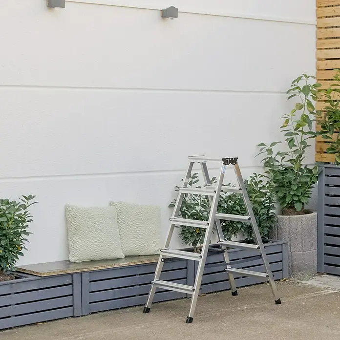 Krause Monto Doppel-Stehleiter Dopplo - Arbeitshöhe: 2,65 m, Anzahl Tritte: 10 Stk., Belastbarkeit: 150 kg, Aluminium - Eine silberne Klapptrittleiter mit fünf Stufen steht auf einer Terrasse. Im Hintergrund sind ein Sitzbereich und Pflanzen zu sehen.