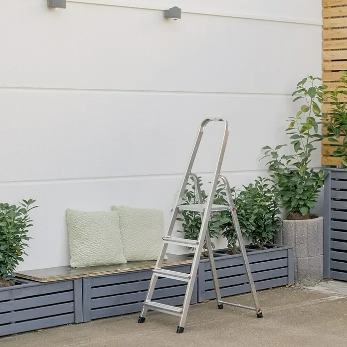 Krause Corda Stehleiter - Arbeitshöhe: 2,8 m, Anzahl Tritte: 4 Stk., Belastbarkeit: 150 kg, Aluminium - Eine silberne Leiter mit vier Stufen steht im Freien vor einer weißen Wand, neben Pflanzen und einem grauen Holzbänkchen mit Kissen.