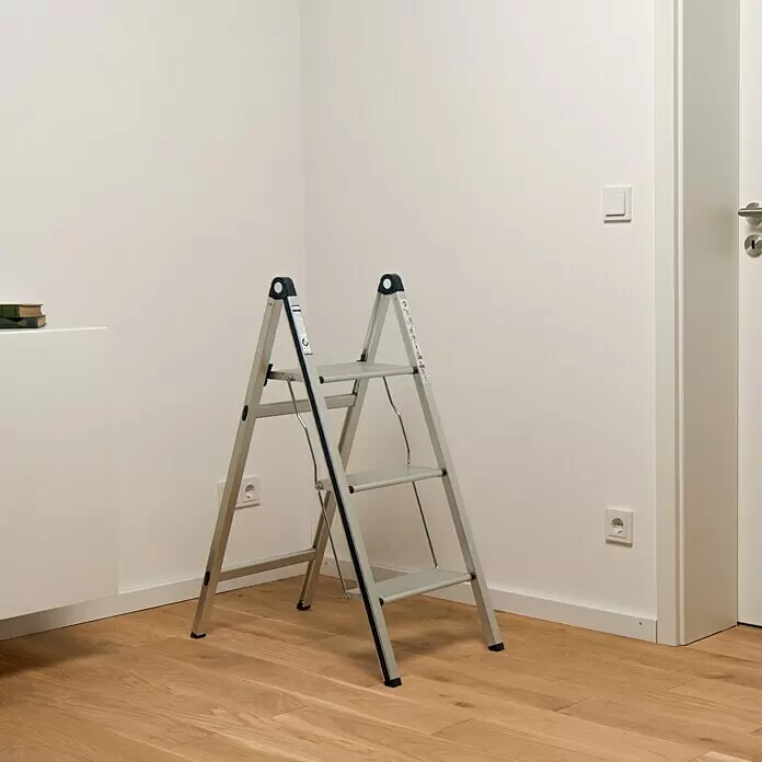 Stabilomat Basicline Klapptritt Slim - Anzahl Tritte: 3 Stk., Arbeitshöhe: 2,75 m, Belastbarkeit: 150 kg, Aluminium - Silberne Aluminium-Klapptrittleiter mit drei Stufen und blauen Akzenten, steht in einem Raum mit weißen Wänden und Holzfußboden.