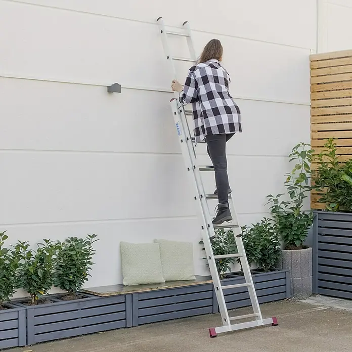 Krause Corda Schiebeleiter - Anzahl Tritte: 16 Stk., Arbeitshöhe: 4,95 m, Belastbarkeit: 150 kg, Aluminium - Eine Frau steigt auf eine Krause Leiter an einer weißen Wand hoch, um Arbeiten zu erledigen. Die Leiter ist stabil und sicher.