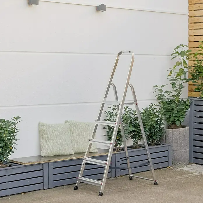Krause Corda StufenstehleiterArbeitshöhe: 3 m, Anzahl Tritte: 5 Stk., Tiefe Stufen: 80 mm, Aluminium - Silberne Haushaltsleiter mit vier Stufen, ideal für Renovierungsarbeiten im Haus und Garten, vor einer weißen Wand und Pflanzen.